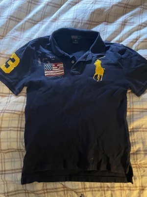 Marinblå piké Polo Ralph Lauren - Snygg marinblå/mörk lila pikétröja från Polo Ralph Lauren med stor gul logga på bröstet, USA-flagga patch och siffran 3 på ärmen. Klassisk krage och korta ärmar. Det är storlek M men i barnstorlek, vilket passar ungefär XS, kanske S.