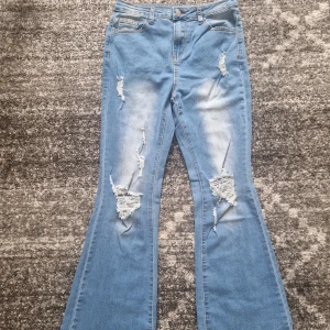 Ljusblå bootcut jeans med slitningar - Säljer ett par ljusblå bootcut jeans med snygga slitningar och rippade detaljer framtill. Jeansen har klassisk femficksdesign, normal midja och är tillverkade i mjukt denimtyg. Perfekta för dig som gillar en avslappnad och trendig look.