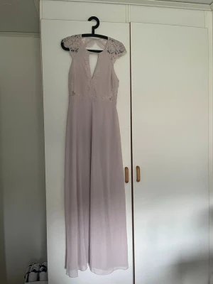 Ljuslila långklänning med spetsdetaljer - Elegant långklänning i ljuslila med spets över axlar och byst, v-ringad hals och öppen detalj framtill. Kjolen är i ett tunt, flytande material som ger ett drömmigt intryck. Perfekt för student eller bal. Den är helt oanvänd och har prislapp kvar!! Hör av er via frågor, prisförslag osv! (Nypris 899kr bevis på bild 4!) 
