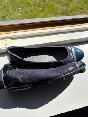 Mörkblå ballerinaskor från Tamaris - Snygga mörkblå ballerinaskor från Tamaris med glansig tå och häl i lackmaterial. Ovandelen är i sammetsliknande tyg med diskreta skiftningar och en dekorativ rem över tån. Skorna har en klassisk rundad form och mjuk innersula för extra komfort.