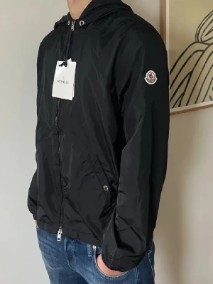 Svart vindjacka från Moncler - Snygg svart vindjacka från Moncler med huva och dragkedja framtill. Jackan har Moncler-logga på ärmen och praktiska fickor med knappstängning. Perfekt för dig som vill ha en stilren och lätt jacka till vardags.