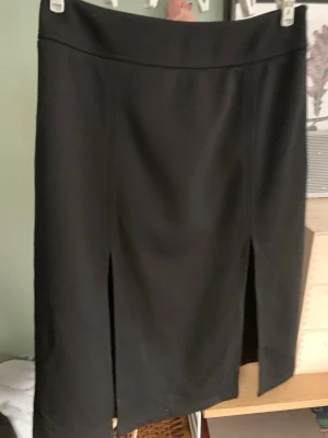 Svart långkjol med slits framtill XS - Stilren svart långkjol med två slitsar framtill. Kjolen har en rak passform och bred linning i midjan. Perfekt för dig som gillar en klassisk och enkel look. Materialet känns mjukt och följsamt.