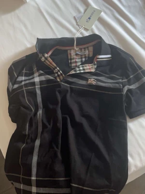 Svart rutig topp från Burberry - Svart topp från Burberry med klassiskt rutigt mönster i grått och vitt samt detaljerad krage med Burberrys signaturrutor. Kortärmad och med liten logga på bröstet. Perfekt för en trendig och stilren look.