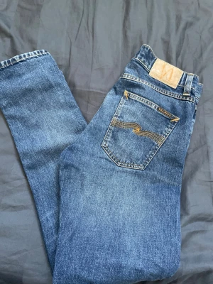 Blå jeans från Nudie Jeans - Snygga blå jeans från Nudie Jeans med klassisk design och gulddetaljer på bakfickan. Jeansen har rak passform och är tillverkade i denim av bomull. Perfekta för en avslappnad och trendig look.