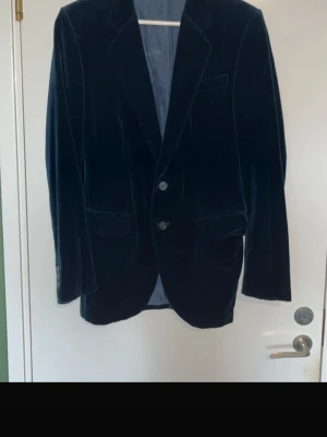 Mörkblå sammetskavaj med två knappar - Stilren mörkblå sammetskavaj med två knappar framtill och klassisk krage. Kavajen har två fickor med lock och en bröstficka. Perfekt för dig som vill ha en elegant och trendig look. Materialet är mjukt och ger en lyxig känsla.