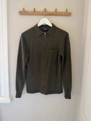 Olivgrön ziptröja från J.Lindeberg - Olivgrön stickad ziptröja från J.Lindeberg med klassisk krage och bröstficka. Tröjan har hel dragkedja framtill och är tillverkad i mjukt material. Perfekt för lager-på-lager och stilren look.