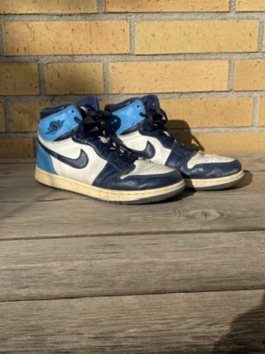 Nike Air Jordan 1 blå och vita sneakers - Snygga Air Jordan 1 sneakers från Nike i blått, vitt och marinblått läder. Klassisk high-top modell med ikoniska Nike logga på sidan och Air Jordan-märket vid ankeln. Skorna har en retro vibe och passar perfekt till streetwear-stilen. Väl använda med en del skav.