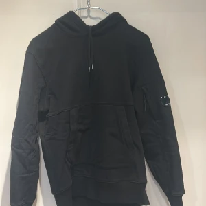 Svart hoodie från C.P. Company - Snygg svart hoodie från C.P. Company med huva och dragsko. Har en unik ficka med dragkedja och linsdetalj på ena ärmen samt stora fickor framtill. Tillverkad i mjukt bomullsmaterial och har ribbade muddar vid ärmslut och nederkant.