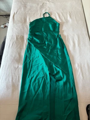 Grön långklänning - Snygg grön långklänning i satinliknande material med en axelbandslös design där banden går runt halsenl. Klänningen har en elegant omlott-detalj framtill och en slank siluett som ger en modern look.