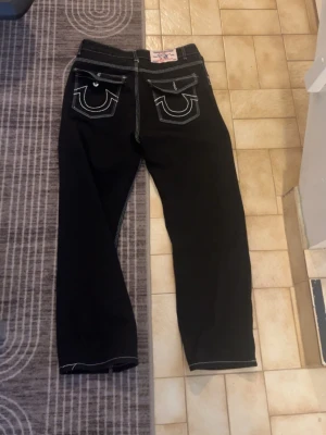 Svarta True Religion jeans  - Svarta jeans från True Religion med raka ben och tydliga vita kontrastsömmar. Klassiska bakfickor med lock och märkets ikoniska brodyr. Jeansen har normal midja och är tillverkade i denim. Perfekta för en cool och avslappnad stil.