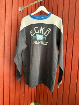Ecko Unltd långärmad tröja grå/blå - Snygg långärmad tröja från Ecko Unltd i grått och blått med vit axelparti och tryck framtill. Tröjan har rund halsringning med blå kant och broderad logga samt texten 'Ecko Unlimited 1972'. Perfekt för en avslappnad streetwear-look.