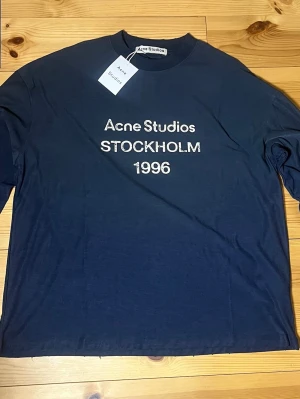 Mörkblå Acne Studios t-shirt - Snygg mörkblå t-shirt från Acne Studios med vit text 'Acne Studios STOCKHOLM 1996' på bröstet. Klassisk rund halsringning och rak passform. Perfekt för dig som gillar stilrena och trendiga plagg med streetkänsla.