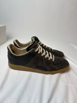 Maison margiela gats - Snygga svarta sneakers med mörkbruna Maison Margielas gats och beige sula. Skorna har vita snören och en klassisk låg siluett. Perfekta för dig som gillar stilrena och tidlösa sneakers med retrovibbar.