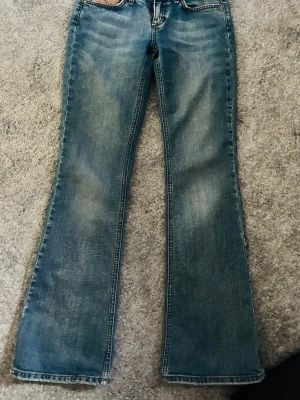 Blå bootcut jeans med färgglada detaljer - Säljer ett par blå bootcut jeans med slitningar och färgglada ränder på bakfickorna. Jeansen har klassisk femficksdesign och kontrastsömmar. Perfekta för dig som gillar en retrostil och vill sticka ut lite extra.