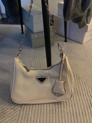 Beige axelremsväska  - Snygg beige handväska med silverfärgad kedjerem och klassisk trekantig logga framtill. Väskan är tillverkad i ett slätt material och har dragkedja upptill samt en dekorativ tag med metallring. Rymmer jätte mycket!🤍