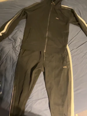 Svarta mjukisbyxor från BOSS - Svarta mjukisbyxor från BOSS med vita sidoremsor längs benen. Byxorna har en sportig look och elastisk midja. Materialet ser ut att vara mjukt och bekvämt, perfekt för en avslappnad stil. Matchar med hoodie i samma design.