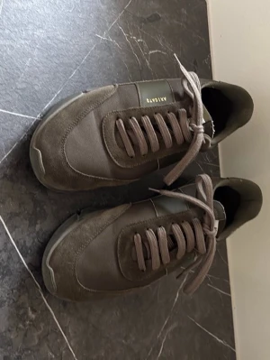 Bruna sneakers från Axel Arigato - Snygga bruna sneakers från Axel Arigato med ovandel i mocka och textil. Skorna har klassisk låg profil, matchande bruna skosnören och diskret logga på sidan. Perfekta för en stilren och avslappnad look. Tillverkade i Portugal. Pris kan diskuteras, hör av dig!