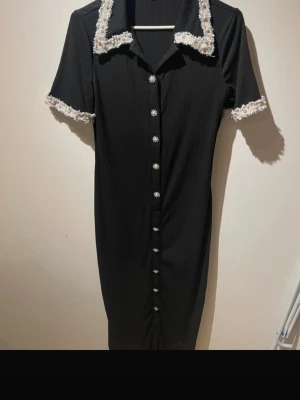 Svart långklänning med pärlknappar - Elegant svart långklänning med korta ärmar och dekorativa pärlknappar framtill. Klänningen har vit spetsdetalj på krage och ärmslut samt en klassisk krage. Materialet är ribbat och ger en snygg struktur. Perfekt för dig som vill ha en chic och tidlös look.