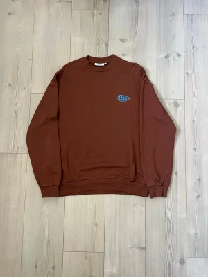 Brun sweatshirt med blått broderi - Säljer en brun sweatshirt från Junkyard med rund hals och blått broderi på bröstet. Tröjan har ribbade muddar vid ärmslut och nederkant. Perfekt för en avslappnad och stilren look.