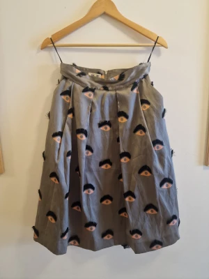 Grå kjol med ögonmönster - Unik grå kjol med broderade ögon i svart och beige. Kjolen har en vid, veckad form och dragkedja bak. Perfekt för dig som vill sticka ut med ett coolt och konstnärligt plagg.
