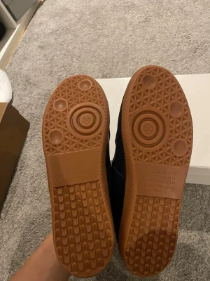 Svarta sneakers Maison Margiela - Säljer ett par svarta sneakers från Maison Margiela med klassisk låg siluett. Skorna har vita snören och en brun gummisula. Ovandelen är i skinn och mocka för en stilren och lyxig känsla. Perfekta för dig som gillar minimalistisk design och premiumkänsla.