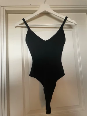 Svart body med smala axelband - Snygg svart body med smala axelband och djup v-ringning. Kroppsnära passform och klassisk design som passar till många olika looks. Materialet ser ut att vara stretchigt och mjukt, perfekt för lager på lager eller som statement-plagg.