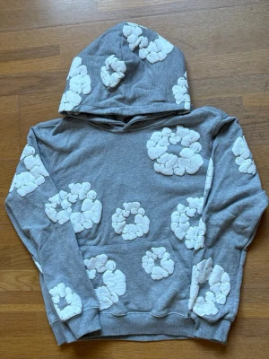 Grå hoodie med vita blommor Denim Tears - Säljer en grå hoodie från Denim Tears med unika vita blommönster över hela tröjan. Hoodien har huva och en klassisk känguruficka framtill. Perfekt för dig som vill sticka ut med en cool och trendig look.