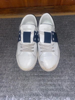 Valentino Garavani Open sneakers vit/blå - Snygga Valentino Garavani Open sneakers i vitt skinn med bred marinblå läderdetalj över mitten. Klassisk rund tå, vita snören och coola nitar på hälen. Perfekta för dig som gillar exklusiva och stilrena sneakers med en twist. Och de kommer med påse 