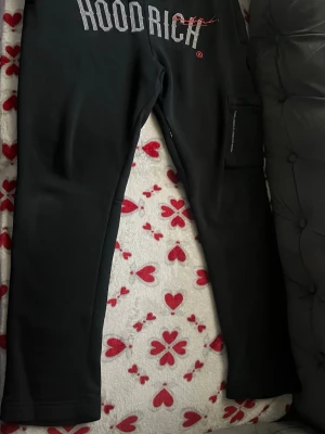 Svarta mjukisbyxor från Hoodrich - Snygga svarta mjukisbyxor från Hoodrich med stort reflekterande tryck på framsidan. Byxorna har en rak passform och en praktisk ficka på sidan. Perfekta för chill dagar eller streetwear-stil.