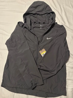 Grå Nike vindjacka  - Snygg grå vindjacka från Nike med huva och dragkedja framtill. Jackan har diskret Nike-logga på bröstet och texten 'NIKE RUNNING' på insidan av huvan. Perfekt för löpning eller som lätt ytterplagg. Stork S men passar också M