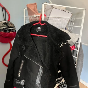 Svart teddyjacka från Zara, storlek L - Säljer en svart teddyjacka från Zara i storlek L. Jackan har en stor krage, detaljer i skinnimitation, dragkedja och bälte nedtill. Fodret är fluffigt och ger en cool look, perfekt för kalla dagar. Snygga metalldetaljer på axeln och vid dragkedjan.