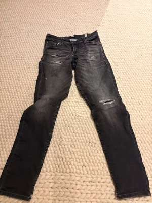 Svarta slitna jeans från Jack & Jones - Säljer ett par svarta jeans från Jack & Jones med slitningar och distressed detaljer på låren och knäna. Jeansen har fickor båda bak och fram , slim passform och klassisk knappgylf. Storlek 158 men passar för personer som är ungefär 166 och neråt eftersom storlekarna är lite konstiga. Lagade själva med lappar men man märker inget från insidan