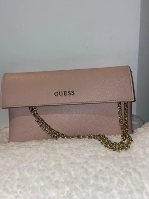 Ljusrosa kuvertväska från Guess - Snygg ljusrosa kuvertväska från Guess med guldfärgad kedja som axelrem. Väskan har stilren design med Guess-logga framtill och är gjord i ett strukturerat konstläder. Perfekt för att lyfta din outfit med en trendig accessoar.