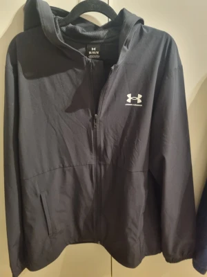 Svart vindjacka från Under Armour - Svart vindjacka från Under Armour med huva och dragkedja framtill. Jackan har två sidofickor och vitt logotryck på bröstet. Tillverkad i lätt och smidigt material med meshfoder för extra komfort. Fast pris 475 kr.