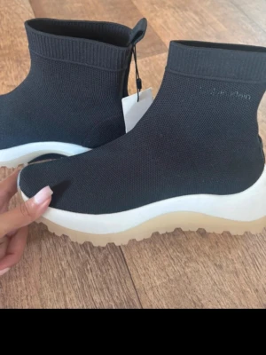 Svarta stövletter från Calvin Klein - Snygga helt nya svarta stövletter från Calvin Klein med stickad ovandel och chunky vit sula. Modellen har en strumpliknande passform och diskret logga på sidan. Perfekta för dig som gillar modern och sportig stil. Kostar runt 2000kr på nätet. Köpta från Kanada. Har tyvärr inte orginal förpackningen kvar.