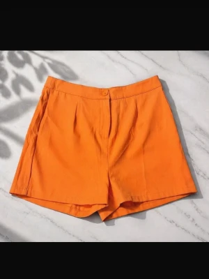 Orange höga shorts i bomull - Snygga orange shorts med hög midja och klassisk knappstängning. De har en enkel design med diskreta veck framtill och är tillverkade i ett mjukt bomullsmaterial som känns bekvämt och luftigt. Perfekta för varma dagar och en färgstark look.