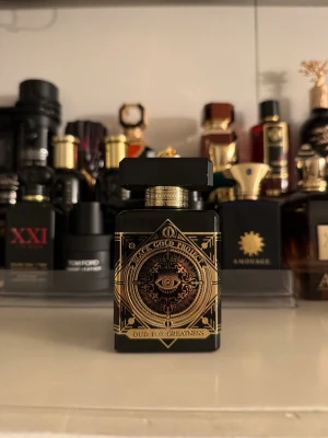 Initio Oud For Greatness parfym - En lyxig parfymflaska i svart med detaljer i guld från Initio Parfums Privés. Flaskan har ett mystiskt öga och geometriska mönster på framsidan. Parfymen heter Oud For Greatness och utstrålar exklusivitet och elegans.