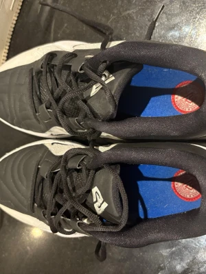 Nike svartvita träningsskor - Säljer ett par snygga träningsskor från Nike i svart och vitt. Skorna har klassisk snörning, vadderad plös och blå innersula med Nike-logga. Ovandelen är i syntetmaterial med struktur för bättre bollkontroll. Perfekta för dig som vill ha stilrena och funktionella skor på planen.