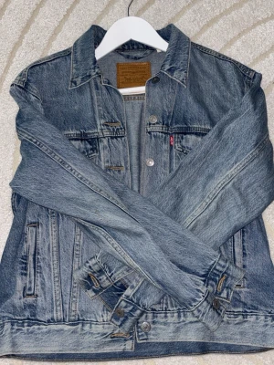 Levi's klassisk blå jeansjacka - Säljer en klassisk blå jeansjacka från Levi's med ikonisk röd logotagg vid bröstfickan. Jackan har två bröstfickor med knapp, sidofickor och silverfärgade knappar framtill. Snygg tvättad denim och tidlös design som alltid funkar.