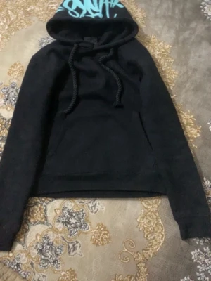 Svart hoodie med blå text på huvan - Säljer en svart hoodie med stor huva och blå textdetalj på huvan. Hoodien har en magficka och tjocka snören vid halsen. Perfekt för en avslappnad och cool stil.