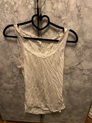 Vit genomskinligt  spetskantad linnetopp från KappAhl - Snygg vit linnetopp från KappAhl med spetsdetaljer längs urringningen och axelbanden. Toppen är ärmlös och har en feminin, enkel design som passar perfekt till jeans eller kjol. Tillverkad i mjuk bomull för en bekväm känsla. Den är rätt så genomskinlig men inte på ett överdrivet sätt 