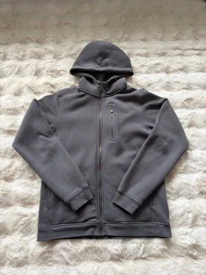 Mörkgrå hoodie med dragkedja - Junior storlek 170. Snygg mörkgrå hoodie med hel dragkedja och huva. Hoodien har långa ärmar och två sidofickor. Perfekt för en avslappnad stil och enkel att matcha med andra plagg. 