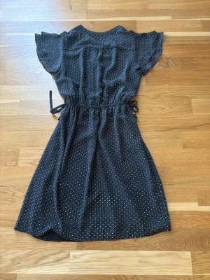 Svart mönstrad klänning  med knytband - Svart klänning med vitt småmönster, rund hals och korta volangärmar. Klänningen har knytband i midjan för en justerbar passform och är tillverkad i ett lätt, mjukt tyg som känns skönt mot huden. Perfekt för dig som gillar en avslappnad men ändå fin stil.Storleken bortklippt men tippar den passar både S och M. Se mått; Axelbredd 35,5 cm Längd 88 cm