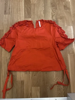Orange tröja - Snygg röd blus med korta ärmar och volangdetaljer längs axlarna. Blusen har en liten v-ringning fram och knytband i sidorna för en cool look. Perfekt för dig som vill sticka ut med färg och detaljer. Materialet känns lätt och luftigt.