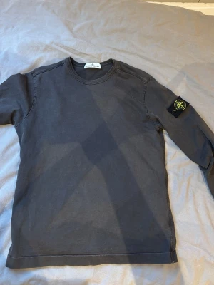 Mörkgrå långärmad Stone Island tröja - Snygg mörkgrå långärmad tröja från Stone Island med den klassiska logotypen på vänster ärm. Tröjan har rund halsringning och är tillverkad i mjuk bomull. Perfekt för dig som gillar streetwear och vill ha en clean look. Går att diskutera priser!