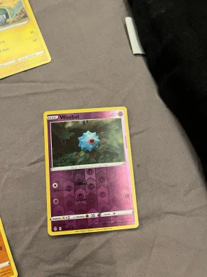 Pokémonkort Woobat holografisk - Säljer ett Pokémonkort med Woobat-motiv. Kortet har en lila och gul ram med holografisk effekt på nedre delen. Woobat är illustrerad i mitten av kortet och det finns tydliga symboler och text. Perfekt för samlare eller Pokémonfans.