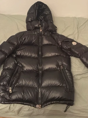 Svart dunjacka från Moncler - Snygg svart dunjacka från Moncler med glansig finish, huva och två dragkedjeförsedda fickor. Jackan har puffig design och Moncler-logga på ärmen. Perfekt för kalla vinterdagar och riktigt varm tack vare dunfyllningen.