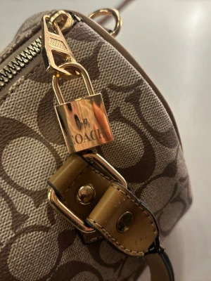 Coach brun monogram handväska - Snygg handväska i beige och brunt monogramtyg med klassiskt C-mönster. Väskan har detaljer i ljusbrunt läder, guldfärgade metalldelar och ett hänglås. Perfekt accessoar för dig som gillar stilrena och trendiga väskor.