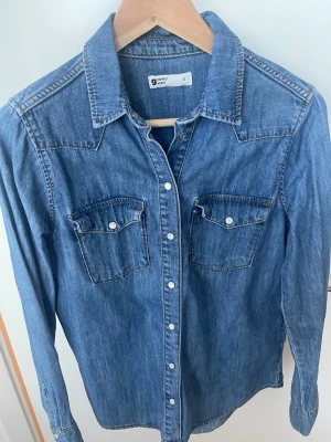  Jeansskjorta. - Snygg blå jeansskjorta  med klassisk westernstil. Skjortan har tryckknappar framtill, två bröstfickor med lock och vit knappdetalj. Tillverkad i mjukt denimtyg och har en markerad krage samt dekorativa sömmar på axlar och rygg.