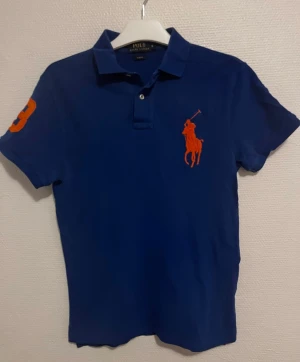 Blå Polo Ralph Lauren piké slim fit S - Klassisk blå pikétröja från Polo Ralph Lauren med orange logga på bröstet och siffra på ärmen. Tröjan har krage, knappar vid halsen och är kortärmad. Perfekt för en sportig och stilren look.